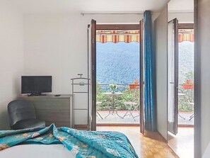 Ein Schlafzimmer mit Bett, Schreibtisch, Stuhl und Fernseher. Es gibt einen Balkon mit Blick auf einen Berg und einen Garten.