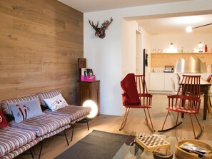 Un salon chaleureux avec un canapé à rayures, un fauteuil rouge, un mur en bois et, au fond, un coin cuisine.