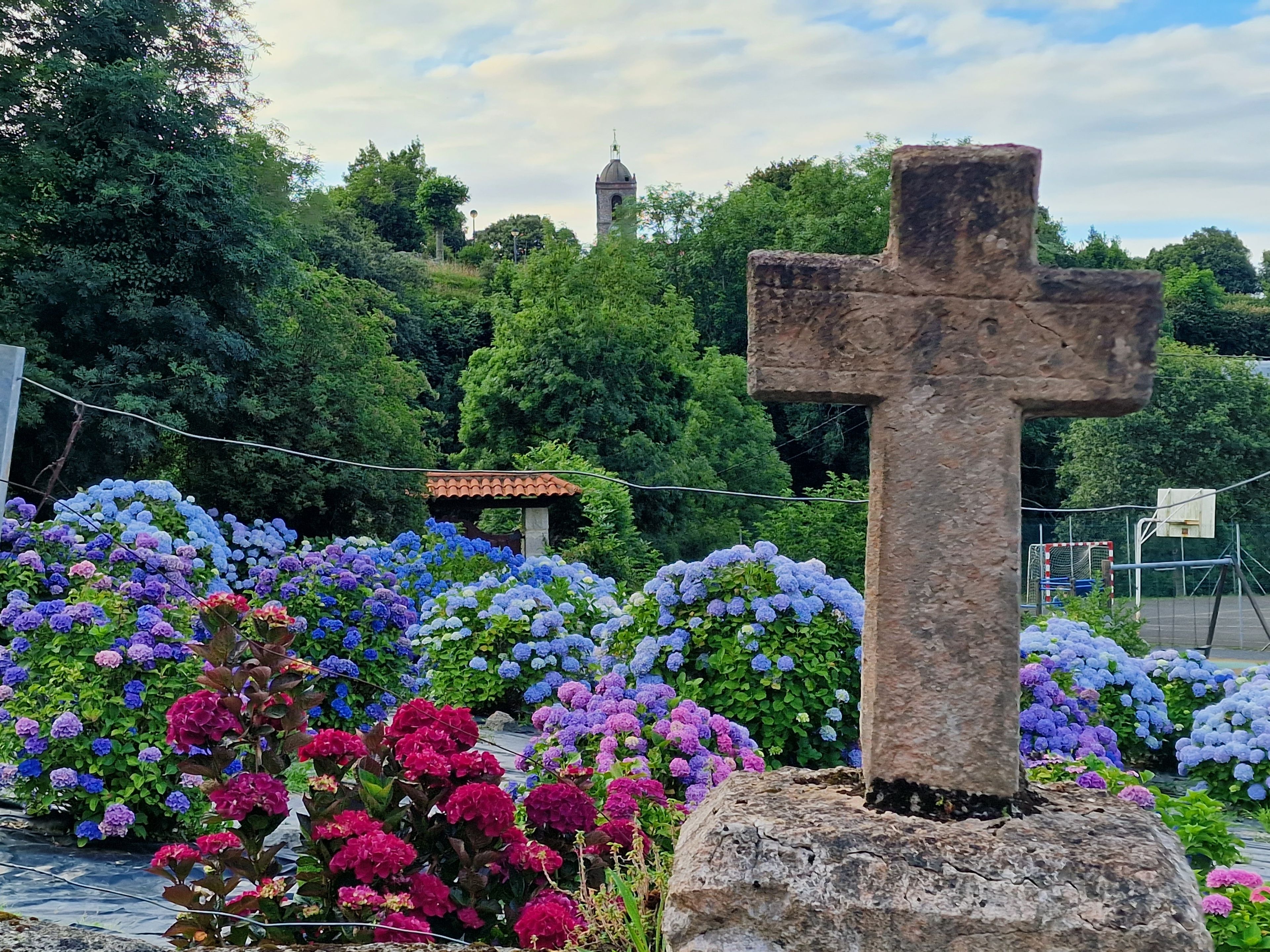 Uma cruz de pedra rodeada por flores vibrantes, com uma torre de sino ao fundo e uma quadra de basquete.