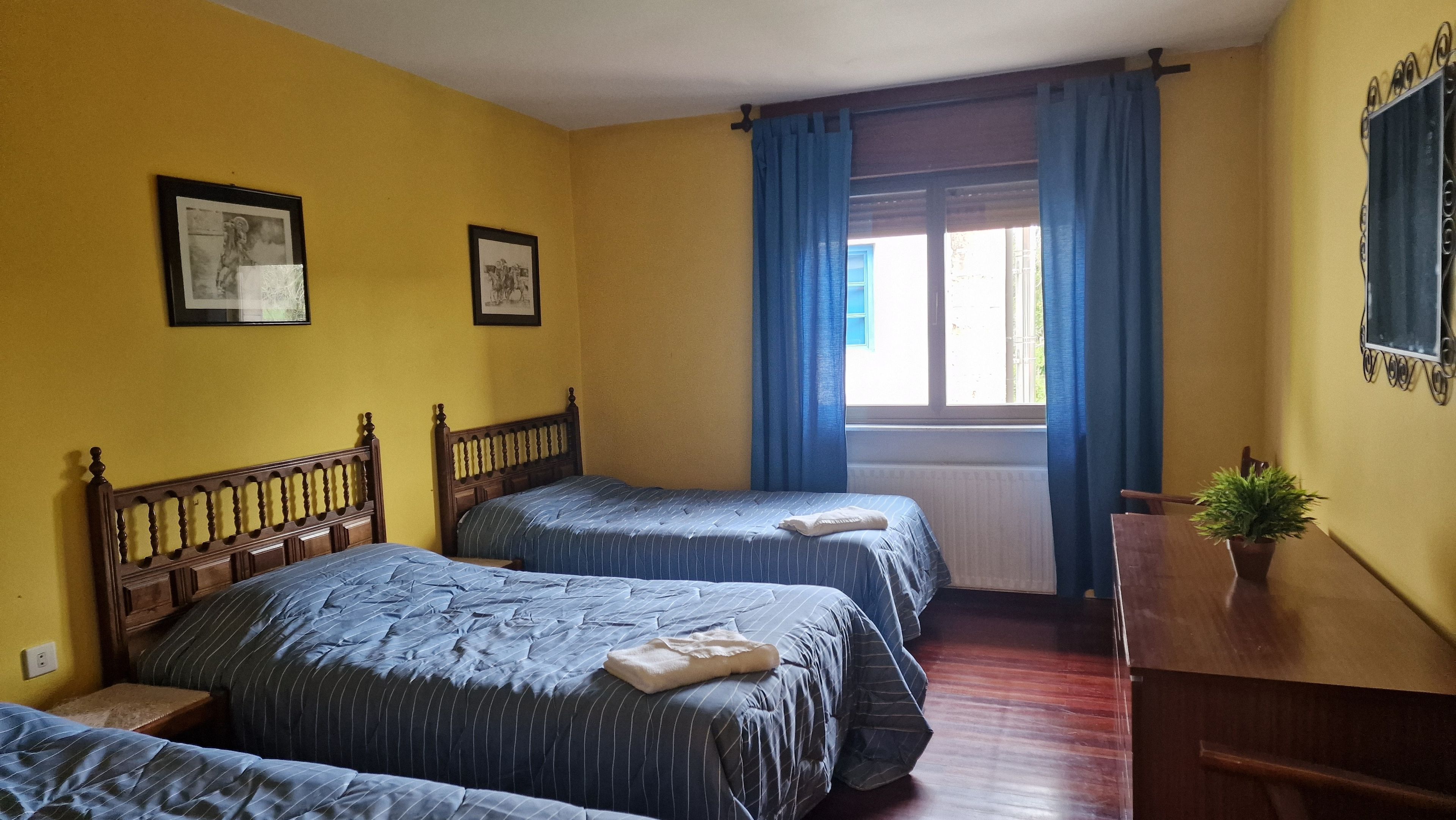 A room with two beds, a wooden headboard, a window with blue curtains, and a small plant on a wooden dresser.