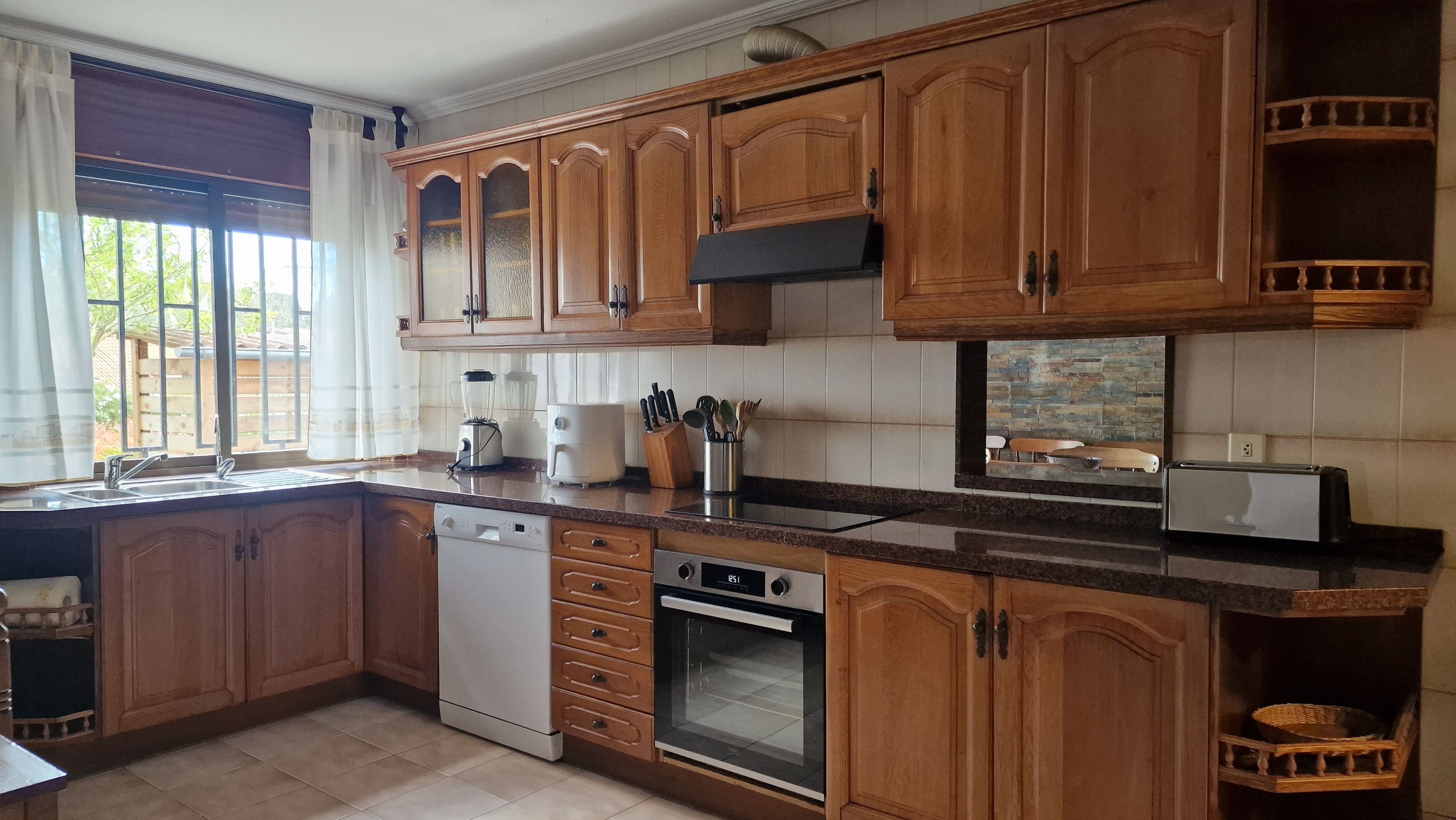 Cozinha com armários de madeira, lava-louças, forno, torradeira e janela com cortinas.
