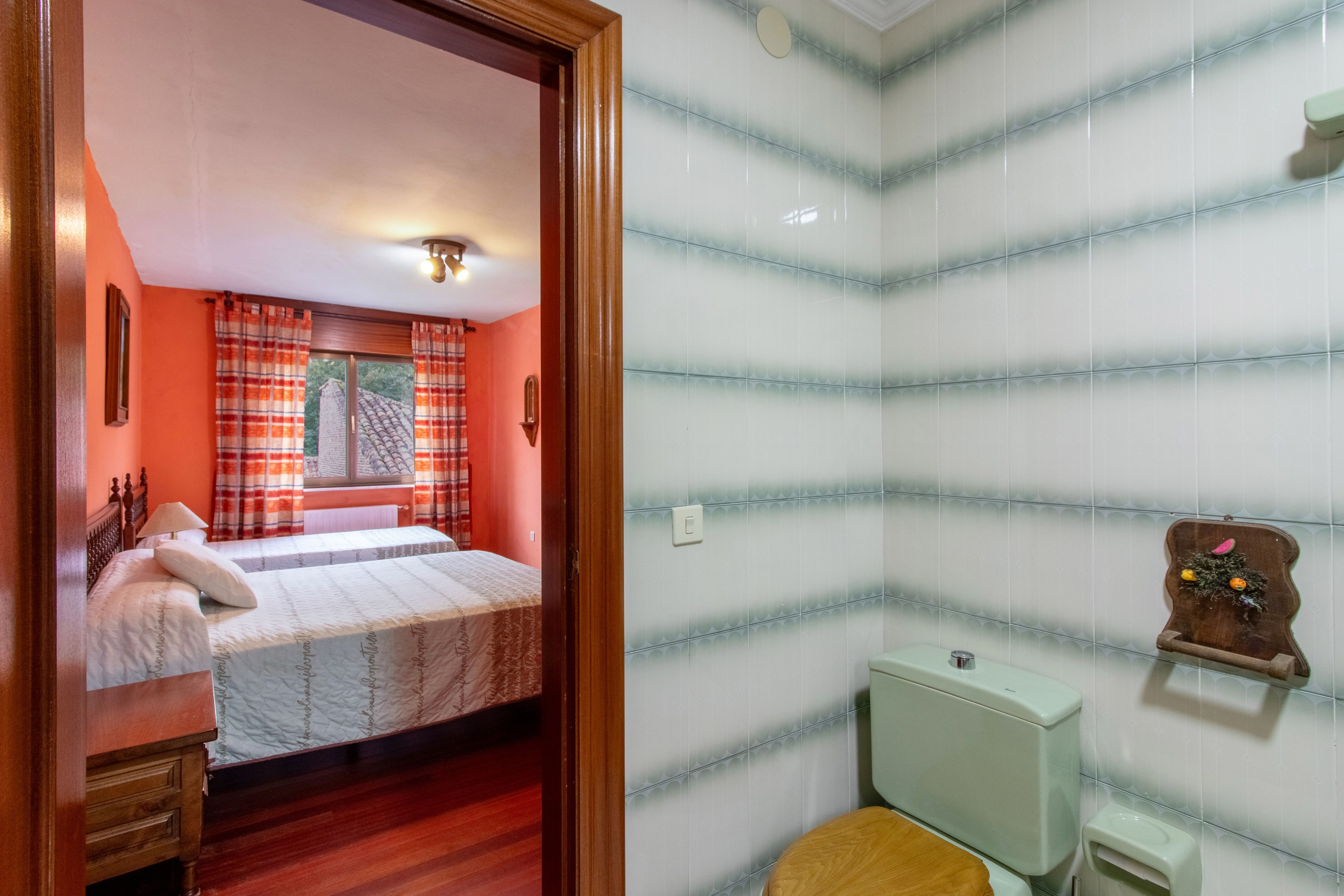 Een slaapkamer met een tweepersoonsbed, een houten nachtkastje, een raam met gordijnen, en een badkamer met toilet en een plank met decoratieve objecten.