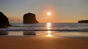Une plage avec un ciel dégagé, une mer calme et un soleil se couchant derrière une formation rocheuse.