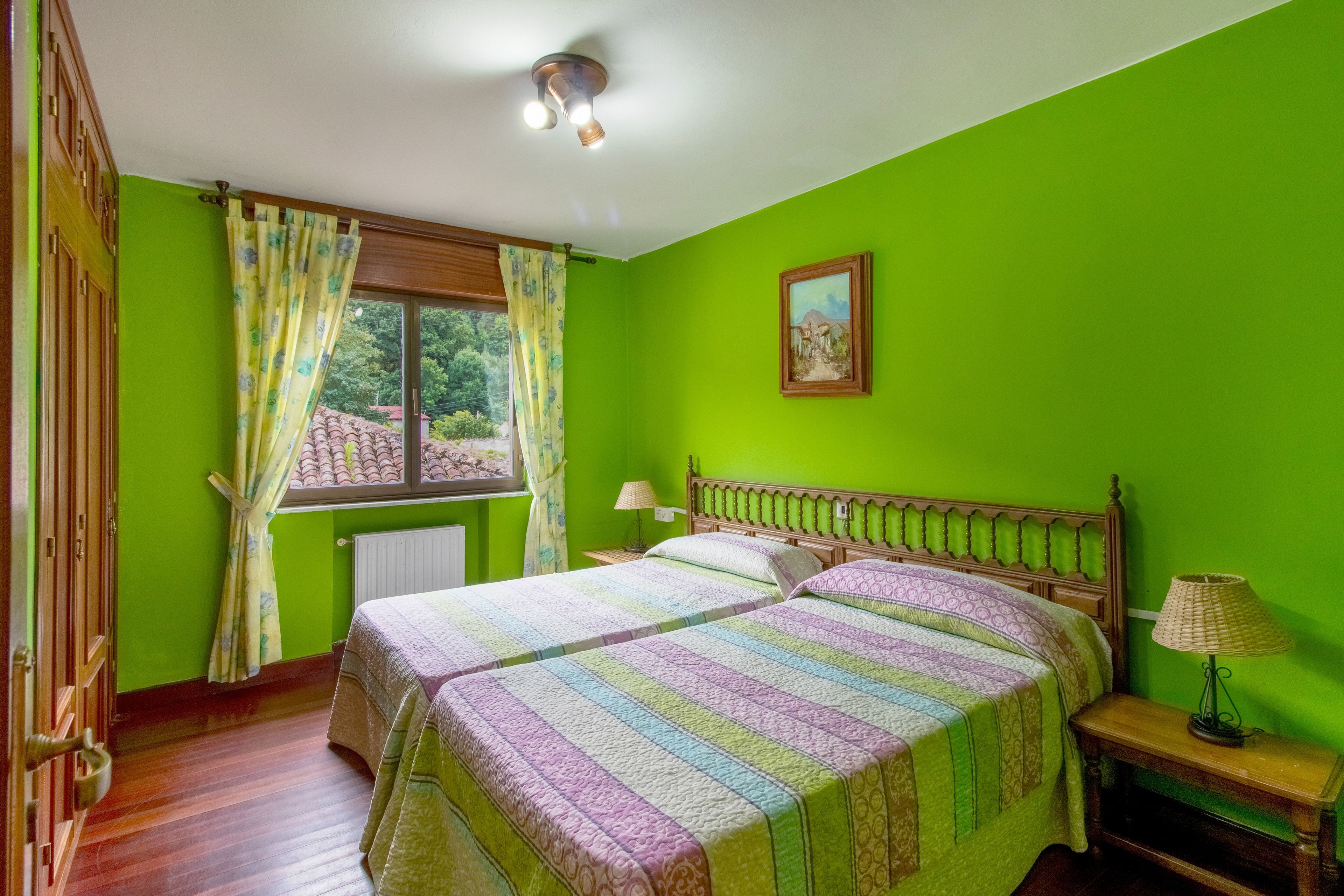 A room with two beds, a window with curtains, a radiator, a lamp, and a painting on the wall.