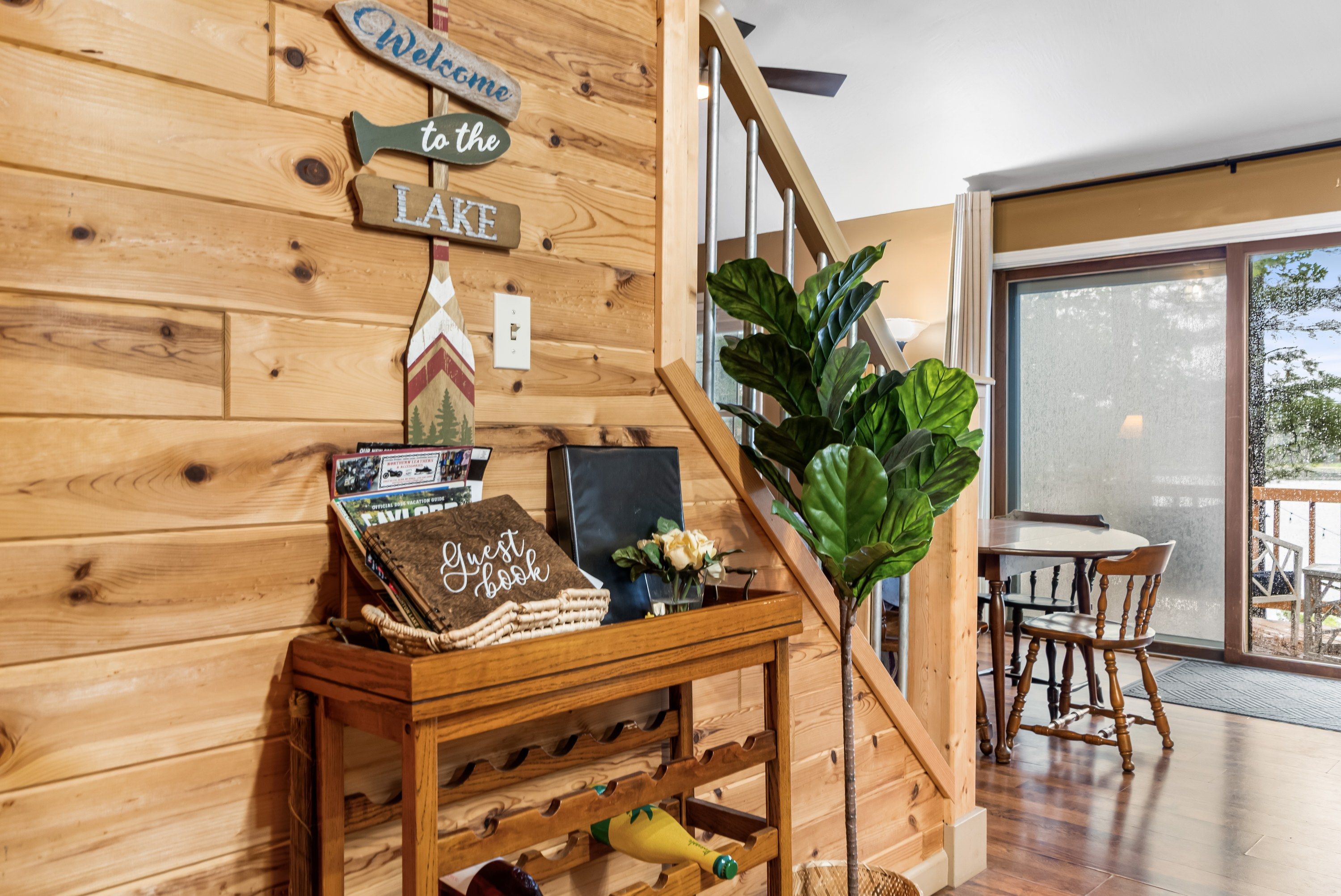 A wooden wall with a welcome sign, a guest book, and a small table with a plant and a bottle.