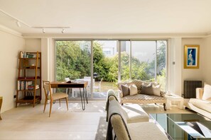 A modern living room with a glass door leading to a garden, a wooden bookshelf, a dining area, and a sofa. - Kensington 2-bed Near Notting Hill With Garden (London)