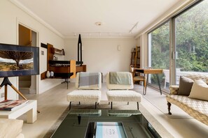 A modern living room with a glass coffee table, two armchairs, a sofa, and a large window offering a view of greenery. - Kensington 2-bed Near Notting Hill With Garden (London)