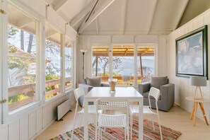 A room with a white table, grey sofas, and a view of a garden and mountains through large windows.