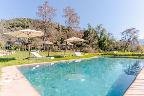 A clear swimming pool with lounge chairs and umbrellas, surrounded by a fenced area with trees and a hill in the background.
