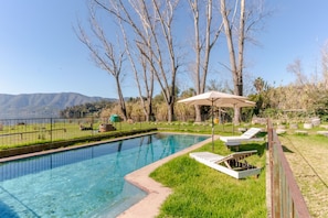 A swimming pool with a view of mountains, surrounded by a grassy area with lounge chairs and an umbrella.