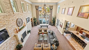 A spacious living room with a fireplace, a large chandelier, and a dining area with a kitchen in the background.