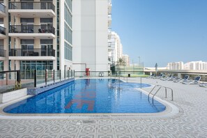 A swimming pool with a tiled edge, surrounded by lounge chairs and a building with balconies. - Stayshort - Peaceful 2-bedroom in Al Furjan That Sleeps 6 (Dubai)