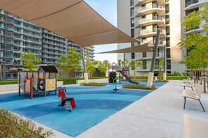 A playground with a slide and swings, surrounded by a building with balconies. - Stayshort - Stunning Beachfront 3-bedroom Apartment Sleeps 5 With Infinity Pool Skyline Views Private Beach Access (Dubai)
