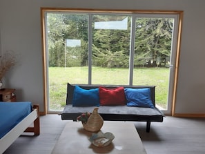 Una habitación con un ventanal grande, un sofá con cojines azules y rojos, una mesita de noche con un jarrón y una cama con cubrecama azul.