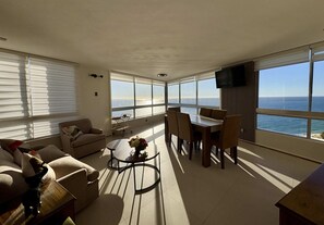 Ein geräumiges Wohnzimmer mit einer Couch, einem Couchtisch und einem Esstisch, große Fenster mit Meerblick und einem flachbildfernseher an der Wand.