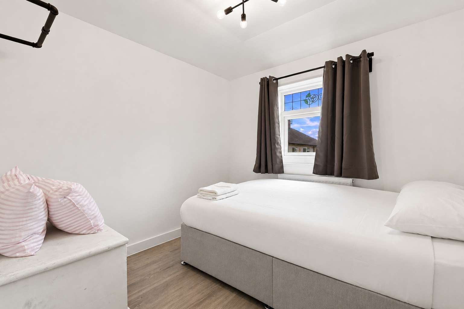 A bedroom with a bed, a window with curtains, a bedside table with pillows, and a ceiling light fixture.