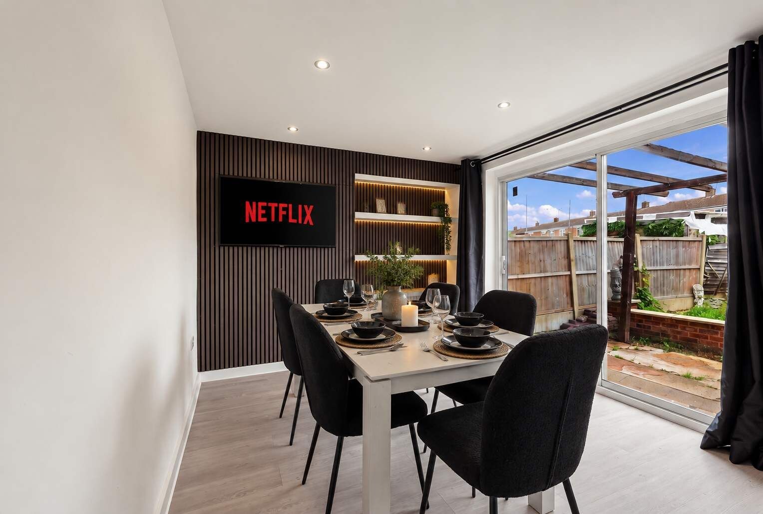 A dining area with a table set for four, a TV mounted on the wall, and a view of the backyard through a glass door.