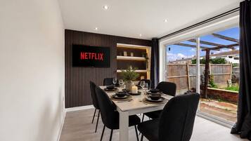 A dining area with a table set for four, a TV mounted on the wall, and a view of the backyard through a glass door.