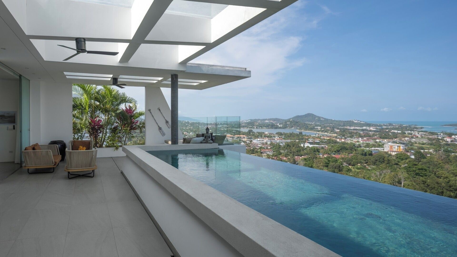 Une piscine à débordement moderne offrant une vue dégagée sur le paysage, un espace salon agrémenté d’une statue de Bouddha et un ventilateur de plafond.