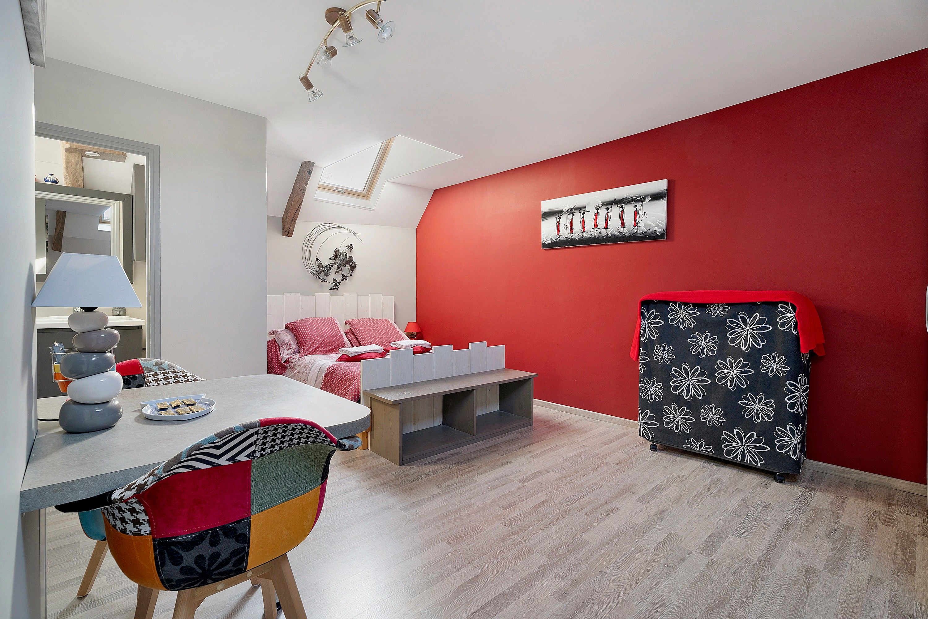 Un dormitorio moderno con una pared de acento roja, una cama con ropa blanca, una mesita de noche, una silla y una obra de arte colgada en la pared.