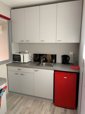Una cocina compacta con armarios blancos, un refrigerador rojo, un microondas, un hervidor y un lavamanos.