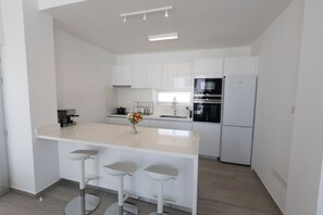 A modern kitchen with white cabinetry, a central island with bar stools, a built-in oven, and a refrigerator. - Luxury 5 Star Cottage – Prime Protaras 1047 (Famagusta)