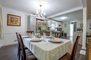 Uma sala de jantar com mesa posta para quatro pessoas, um candelabro e uma cozinha ao fundo.