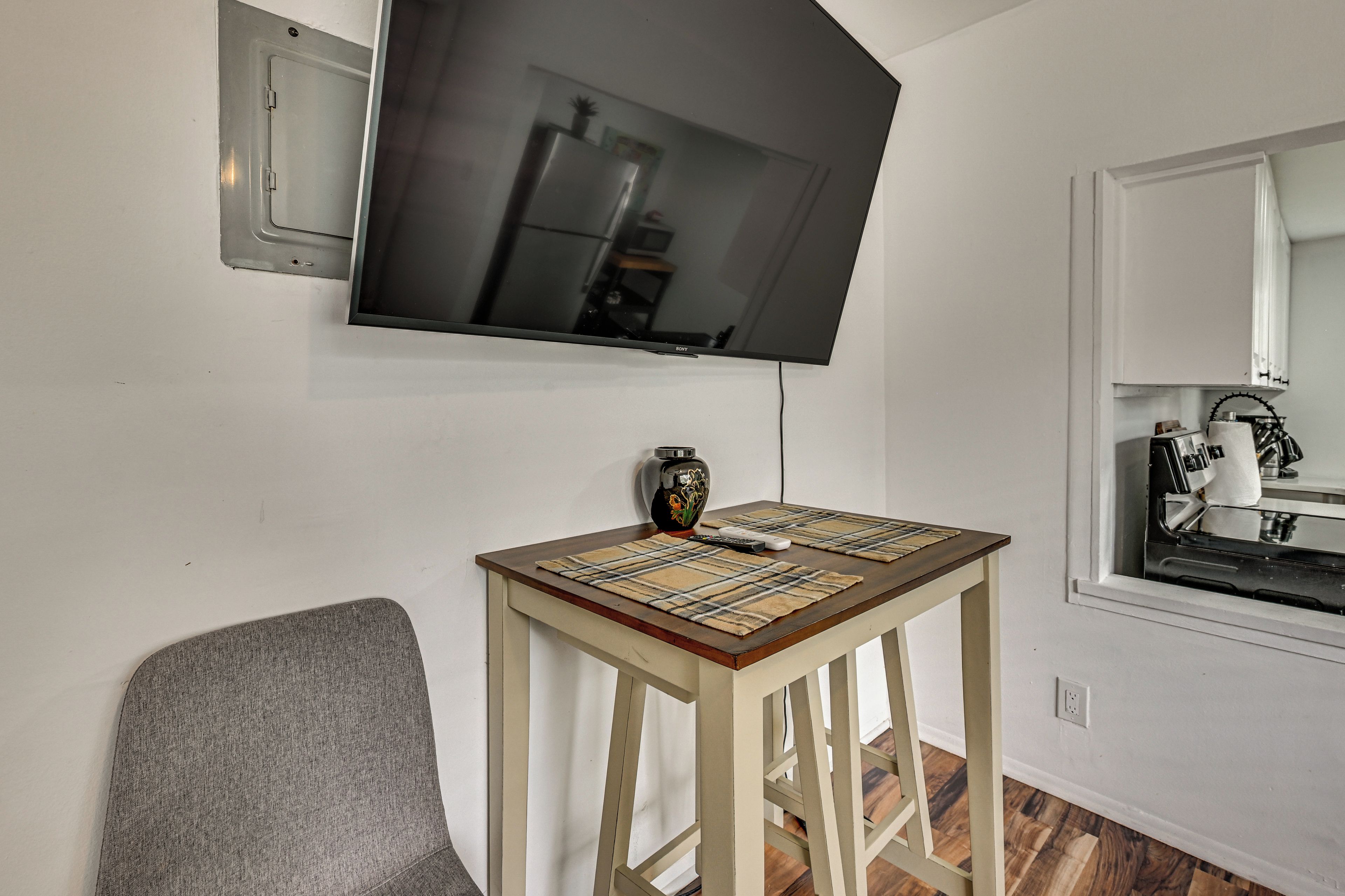 A room with a television mounted on the wall, a small table with a placemat, a chair, and a kitchen area with a stove and a kettle.