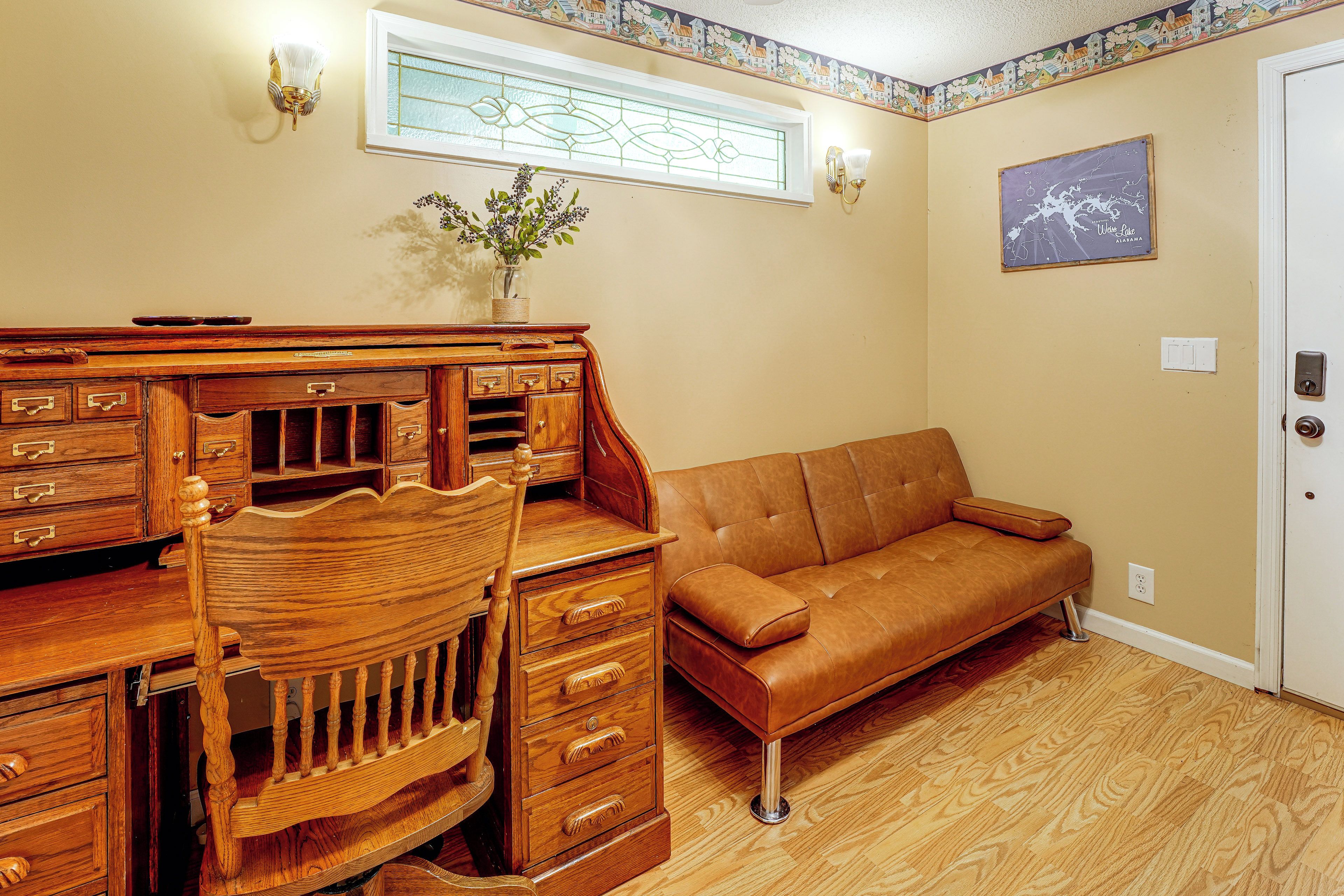 A room with a wooden desk, a leather sofa, a vase with flowers, a stained glass window, and a map on the wall.