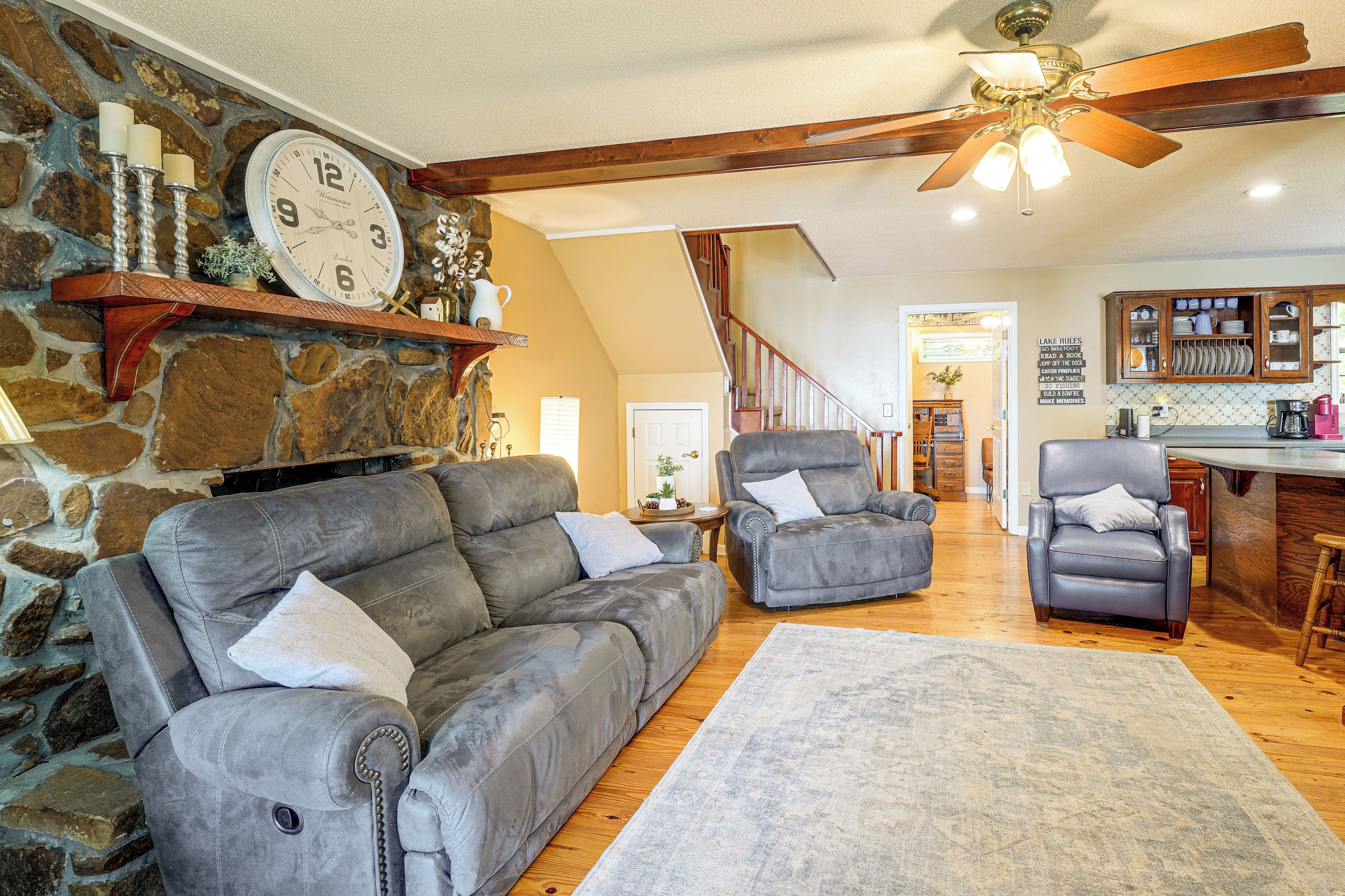 A cozy living room with a stone wall, a fireplace, a ceiling fan, and a kitchen area with wooden cabinets.