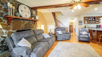 A cozy living room with a stone wall, a fireplace, a ceiling fan, and a kitchen area with wooden cabinets.