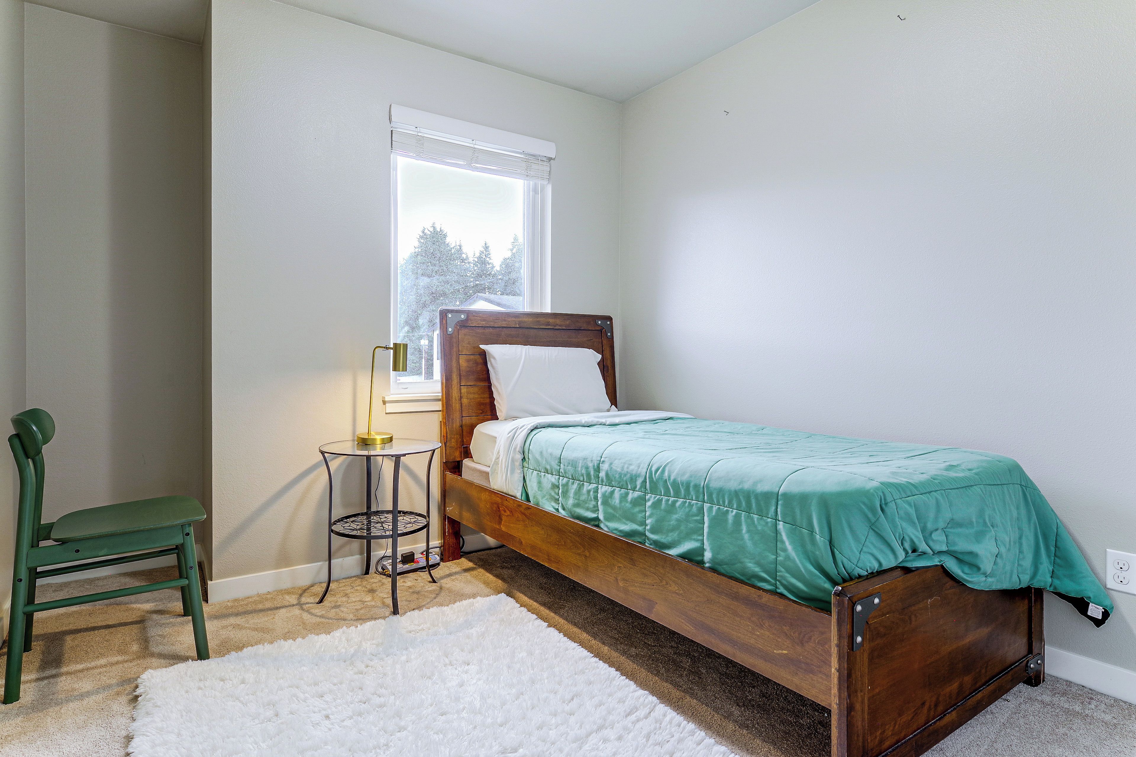 A bedroom with a wooden bed, a green chair, a small round table, and a window with blinds.