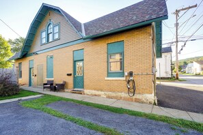 A two-story brick building with a green door and windows, a small porch with chairs, and a visible electrical box on the side.