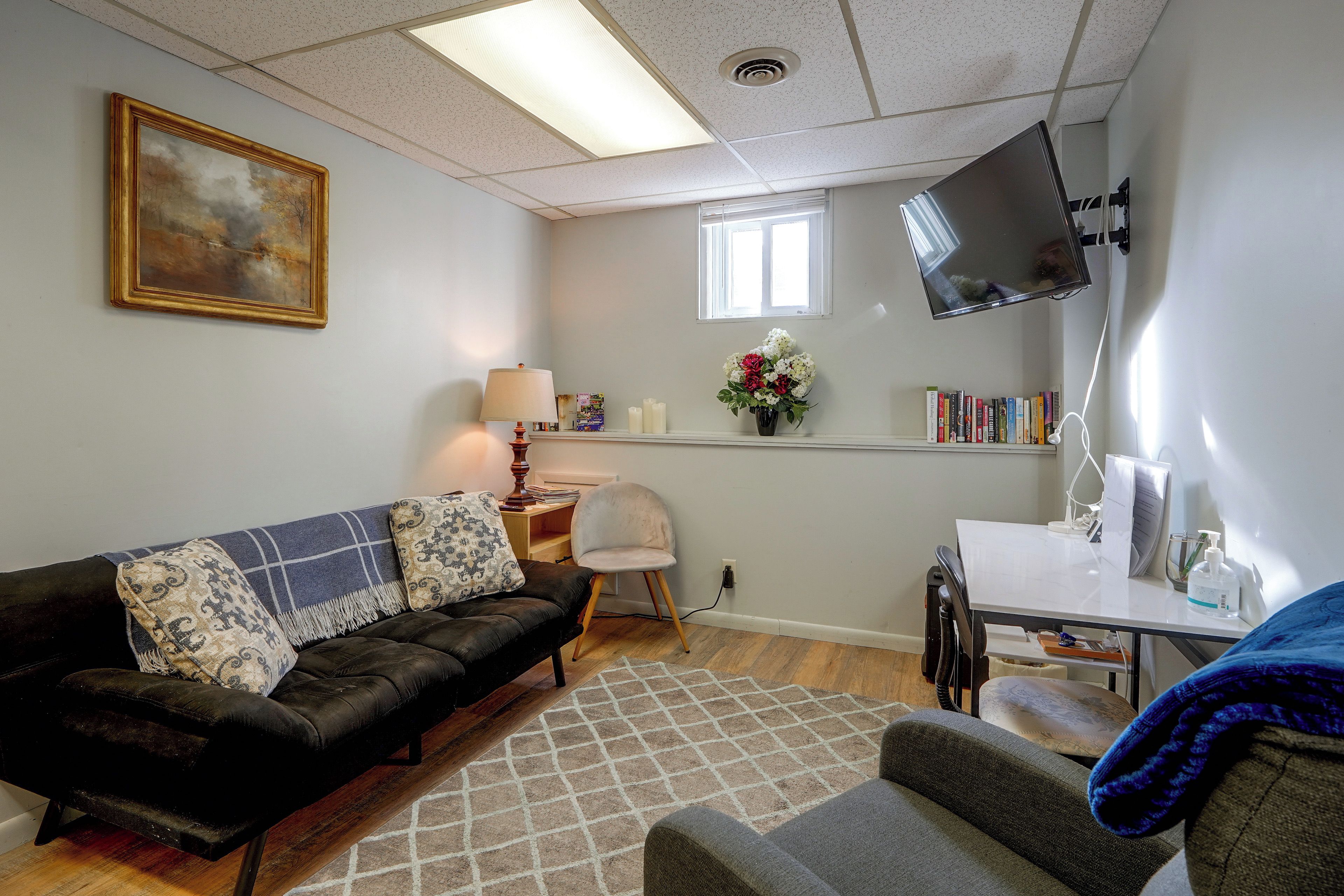 Ein Wohnzimmer mit einem schwarzen Ledersofa, einem kleinen Tisch mit Lampe, einem an der Wand montierten Fernseher und einem Bücherregal mit Büchern.