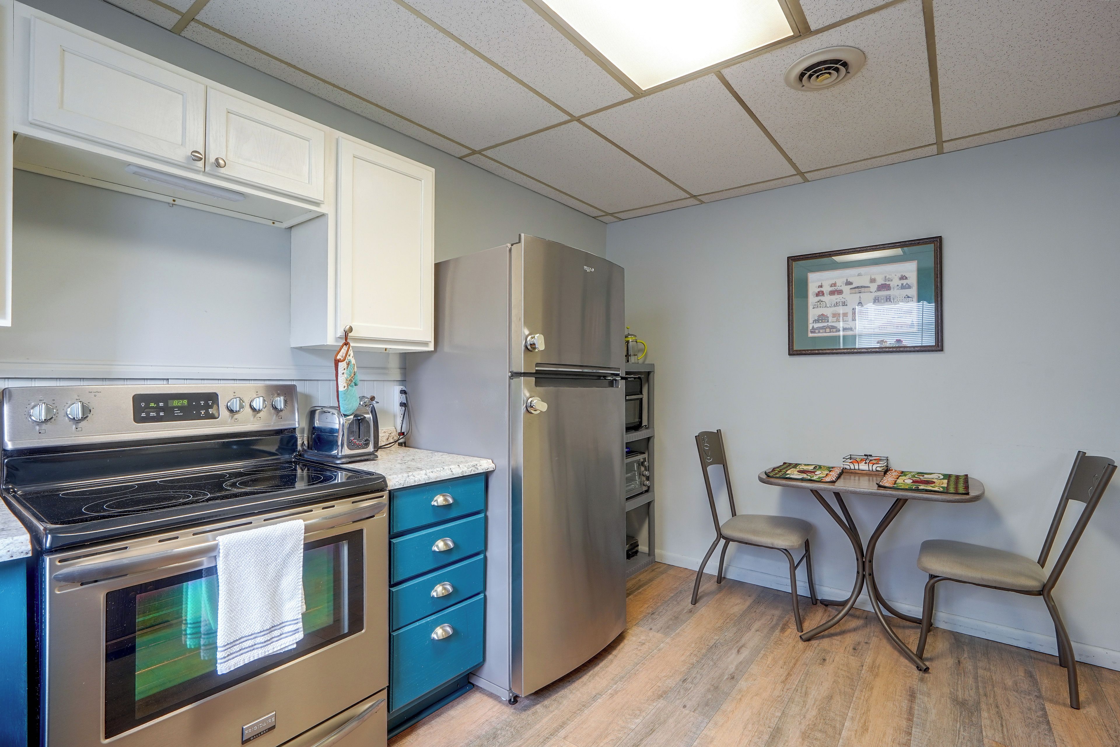 A compact kitchen with a stove, oven, microwave, and refrigerator. There is a small dining table with chairs and a framed picture on the wall.