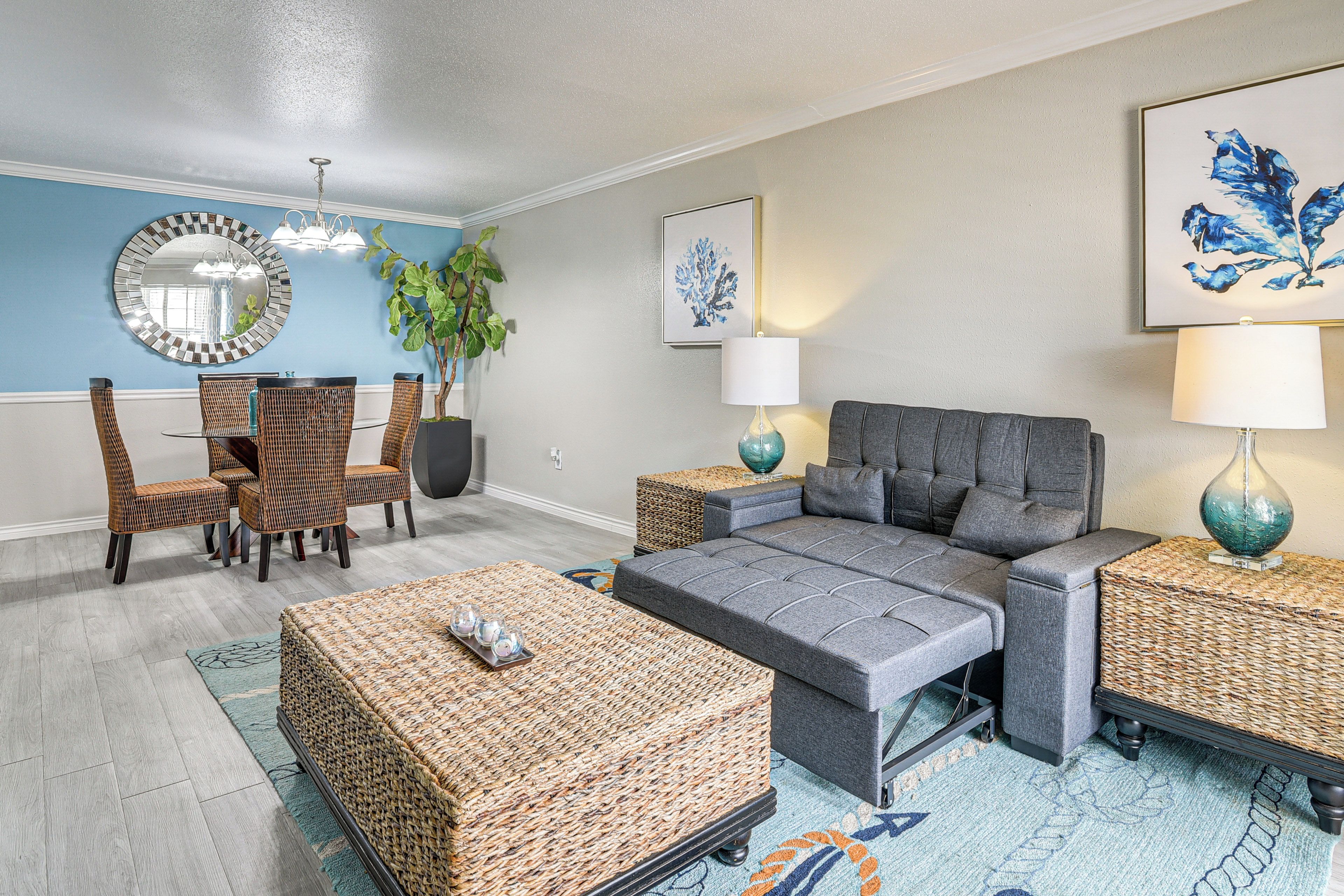 A modern living room with a grey sofa, wicker furniture, a dining area with chairs, and a large mirror on the wall.