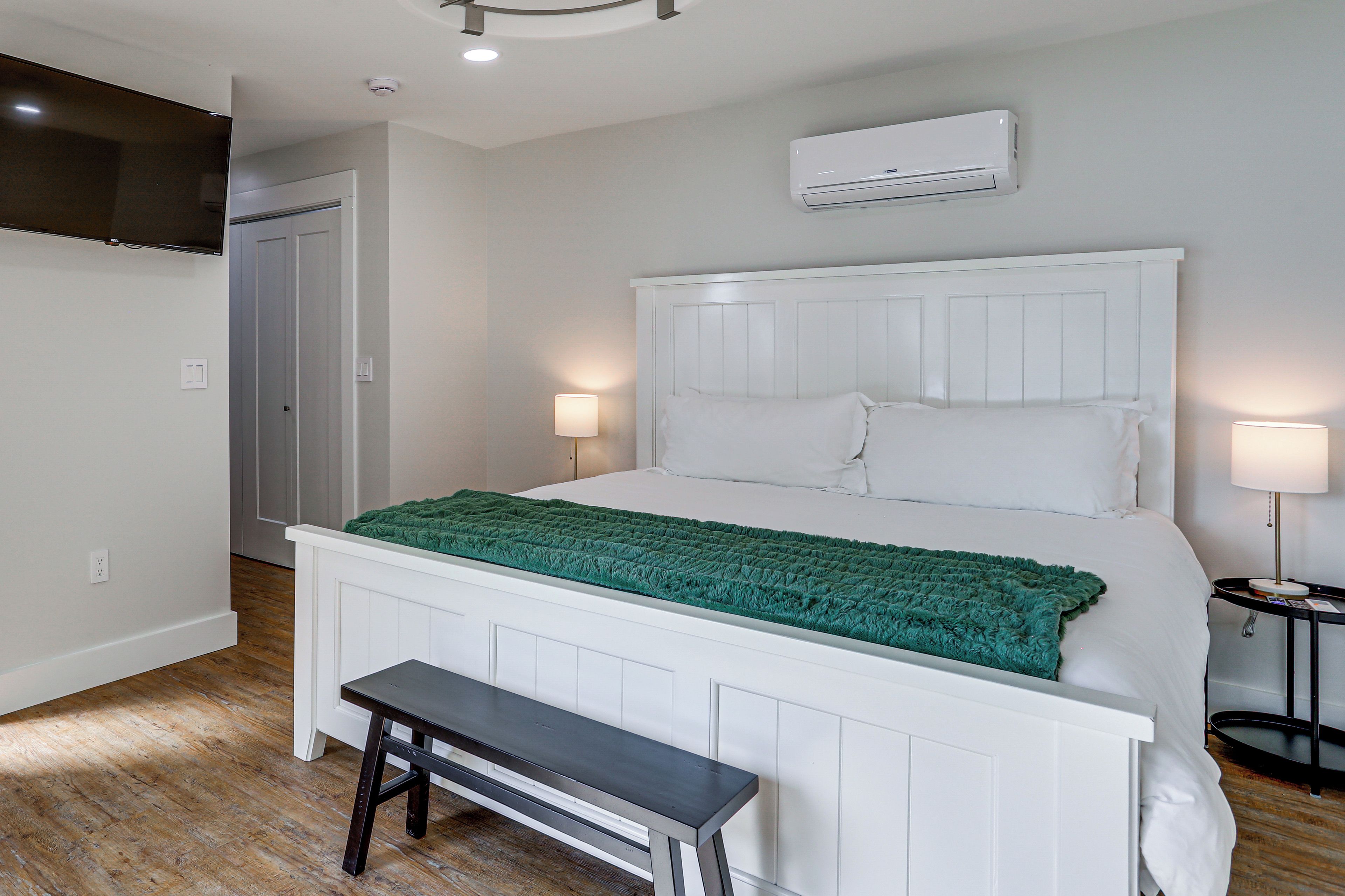A bedroom with a bed, bedside lamps, a bench, an air conditioning unit, and a door.