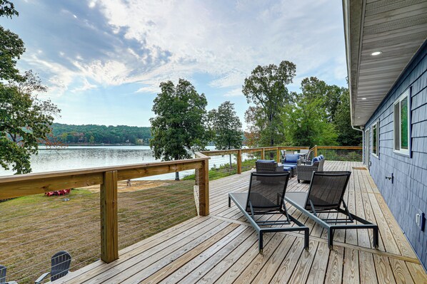 A deck with outdoor furniture overlooking a lake, a wooden railing, and a view of trees and hills in the distance.