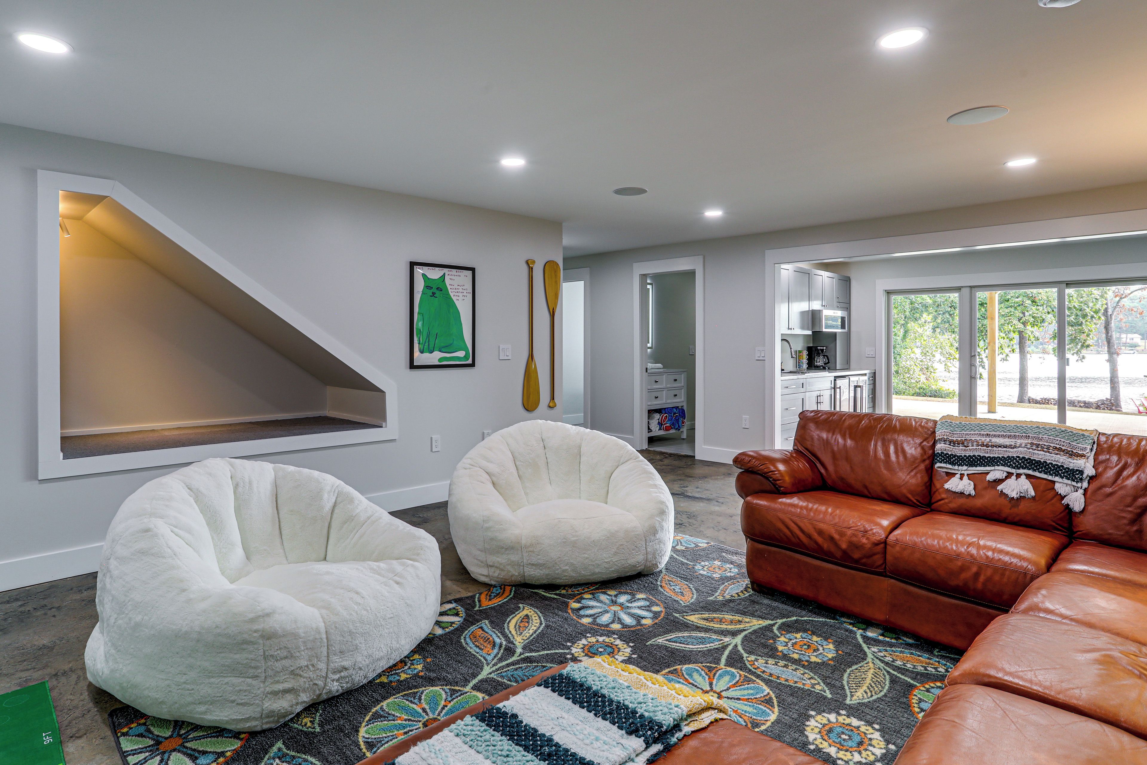 Un salon moderne avec un canapé en cuir marron, deux fauteuils en forme de poire blancs, un tapis coloré et un tableau représentant un chat accroché au mur.