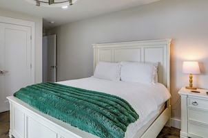 Cama bem arrumada com manta verde, cabeceira branca e mesa de cabeceira com abajur.