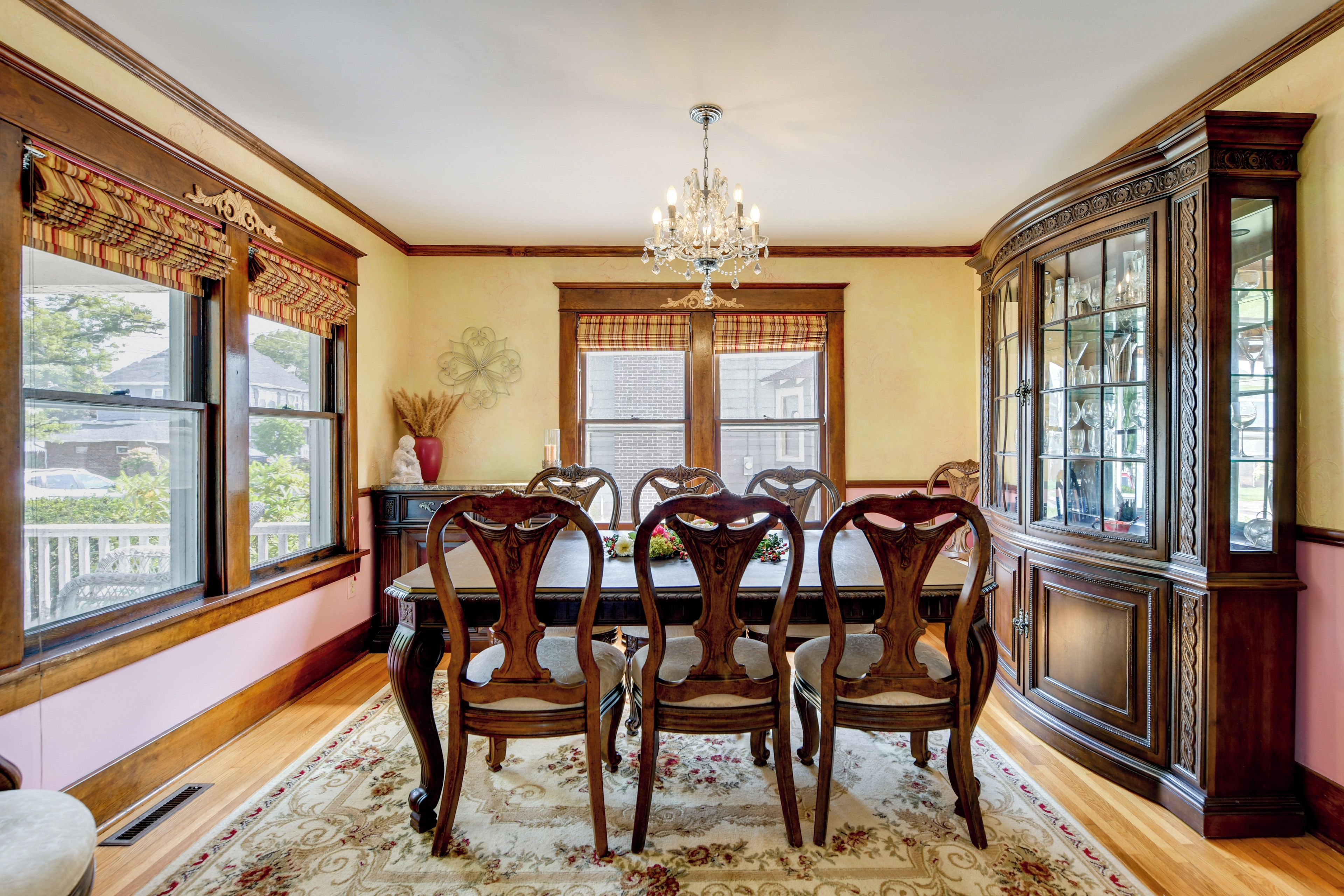 A dining room with a chandelier, wooden chairs, a floral rug, and a china cabinet.