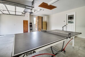 Una mesa de ping pong con red, una puerta de madera y una ventana en una habitación con paredes blancas.