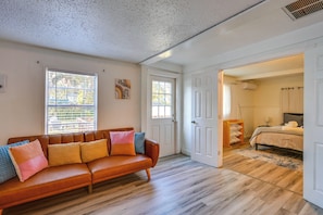 Una habitación con un sofá de cuero marrón, piso de madera y una puerta que da a un dormitorio con cama y mesita de noche.