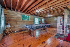 A log cabin interior with a wooden ceiling, a bed, a table, and a painting on the wall.