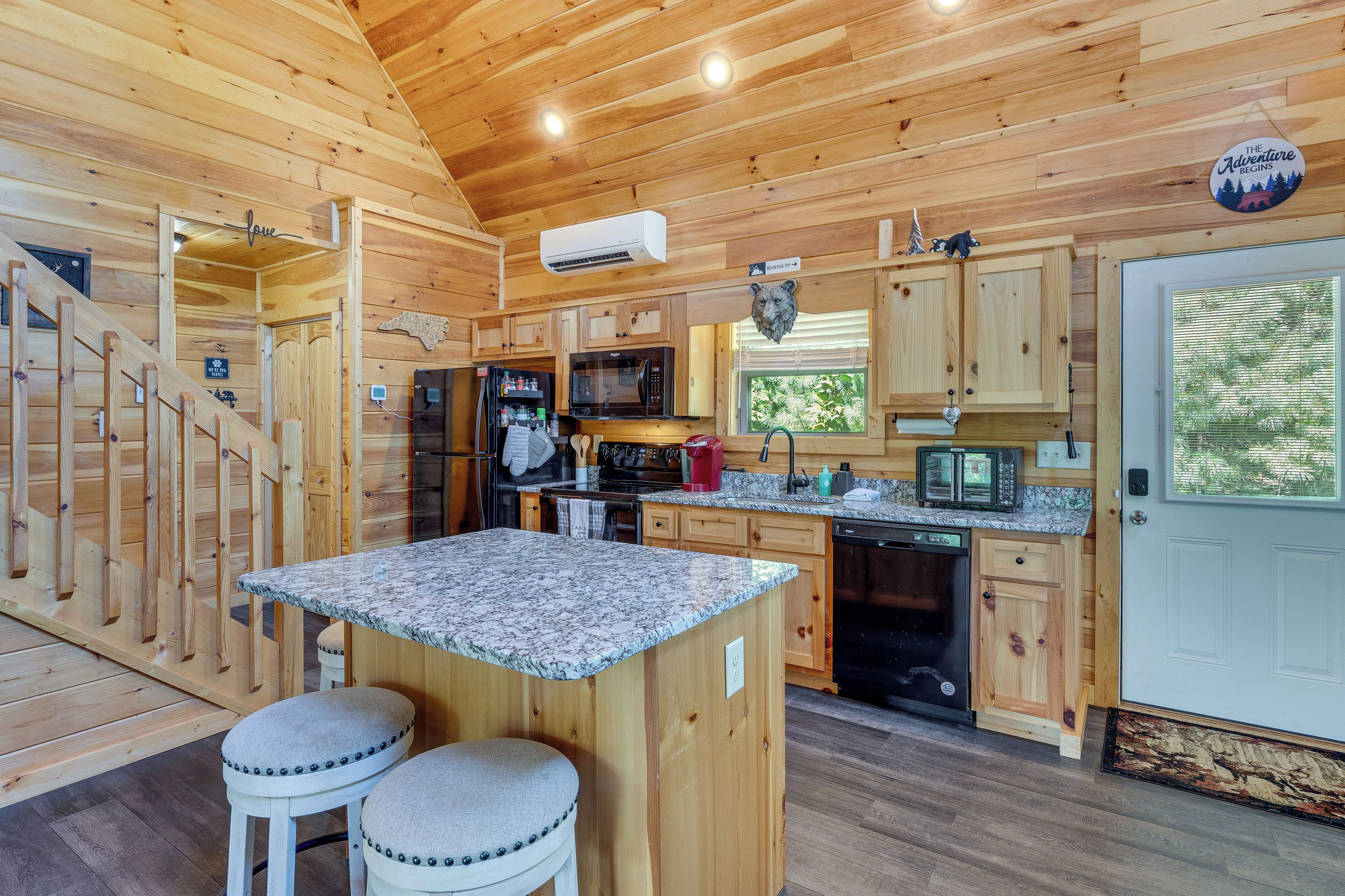 A wooden cabin interior with a kitchen, a dining area with stools, and a staircase.