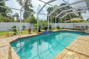 Una piscina con agua cristalina azul, rodeada por una cerca blanca, y un área cubierta con sillas y una sombrilla.
