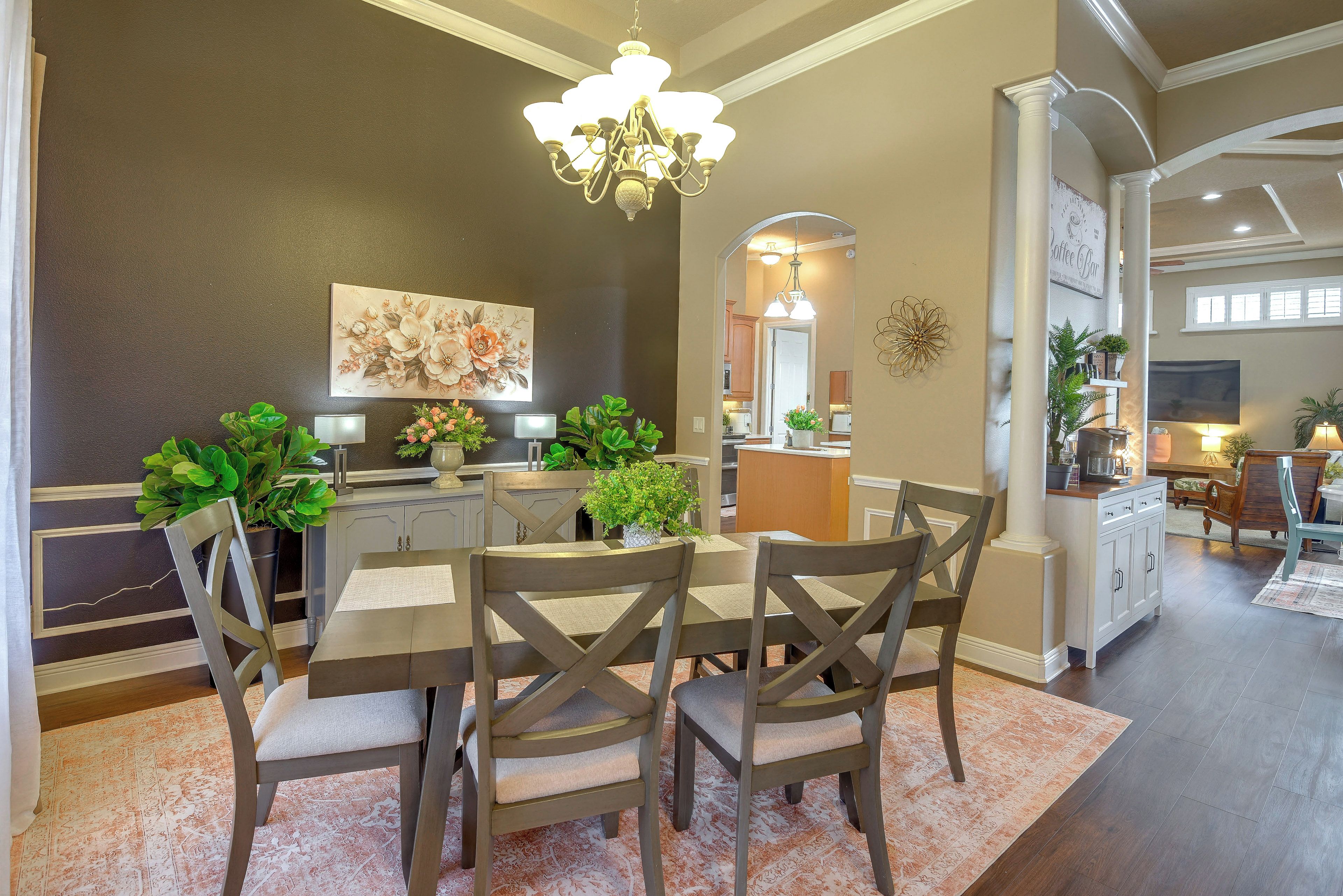 A dining area with a chandelier, a floral painting, and a kitchen in the background.