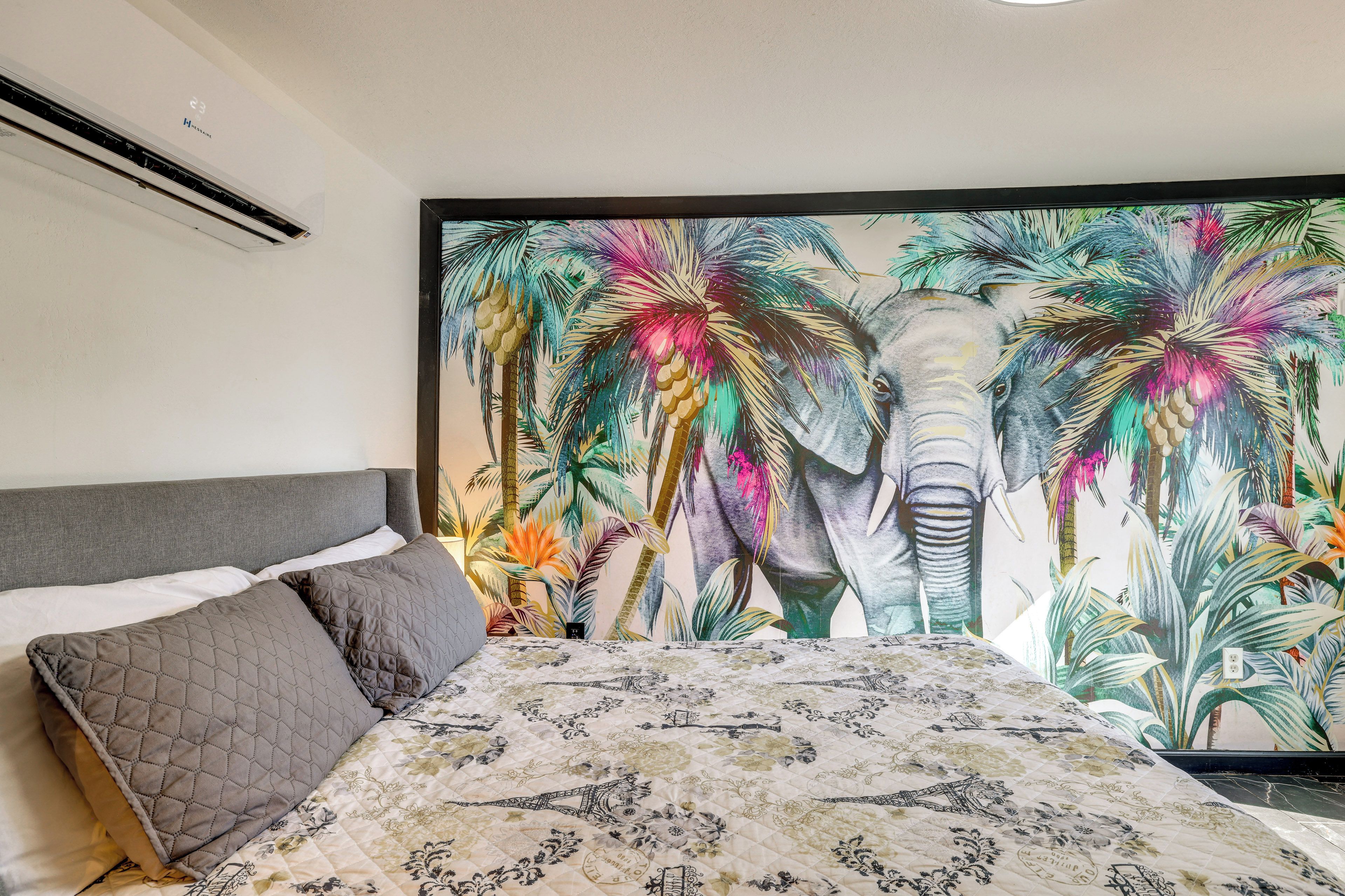 Un dormitorio con una cama grande, un cabecero gris y un mural de elefantes de colores en la pared.