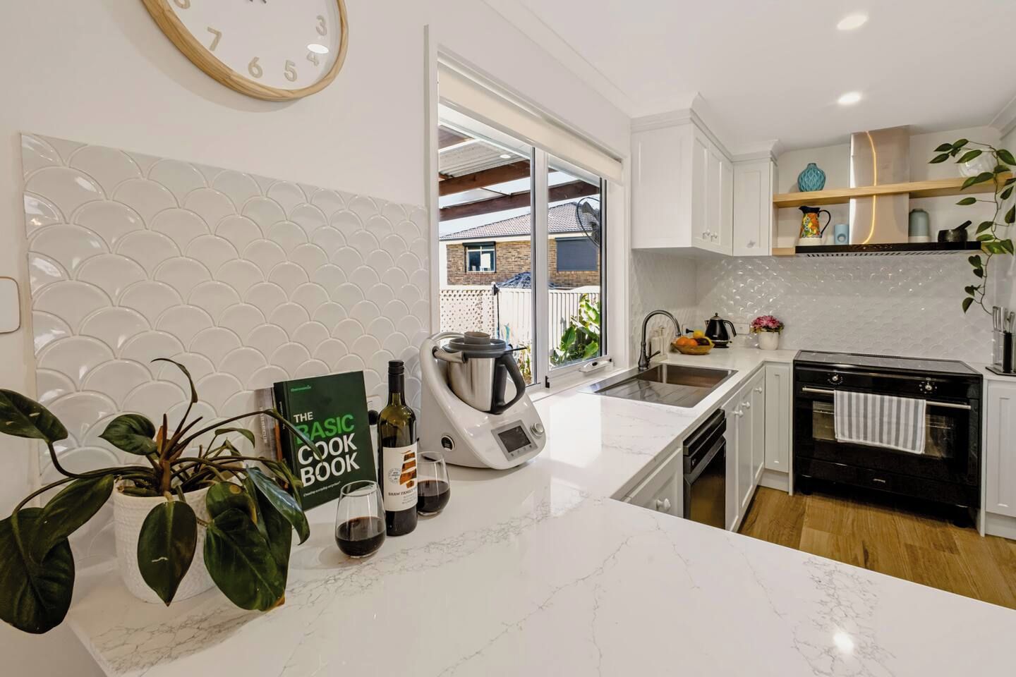 A modern kitchen with white cabinetry, a marble countertop, a kettle, a wine bottle, and a cookbook.