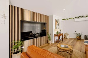 A modern living room with a flat-screen TV mounted on a wooden slat wall, a leather sofa, a small round coffee table, and a potted plant. - Casa Aqua: Tropical/Coastal Escape & McLaren Vale (Christies Beach)